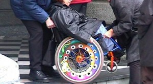 Das Bild zeigt, wie zwei Personen einer Rollstuhlfahrerin über eine Stufe helfen